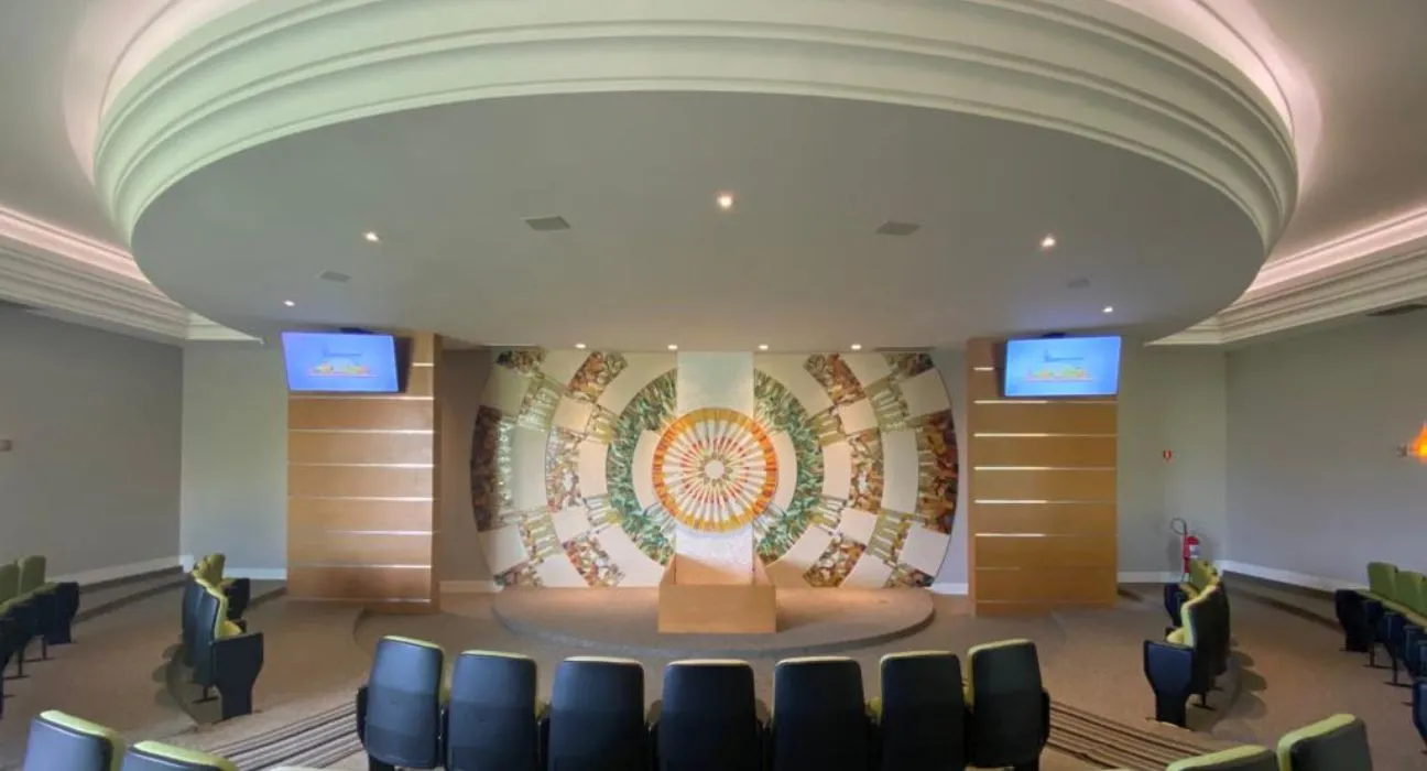 Interior de uma sala com cadeiras dispostas em arco, uma parede decorativa ao fundo e telas de apresentação.