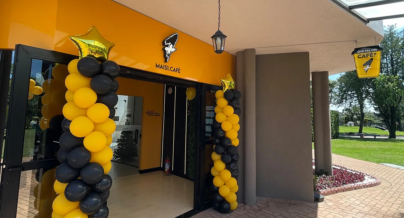 Fachada do Mais Café, com balões amarelos e pretos nas laterais da entrada e uma placa pendurada.