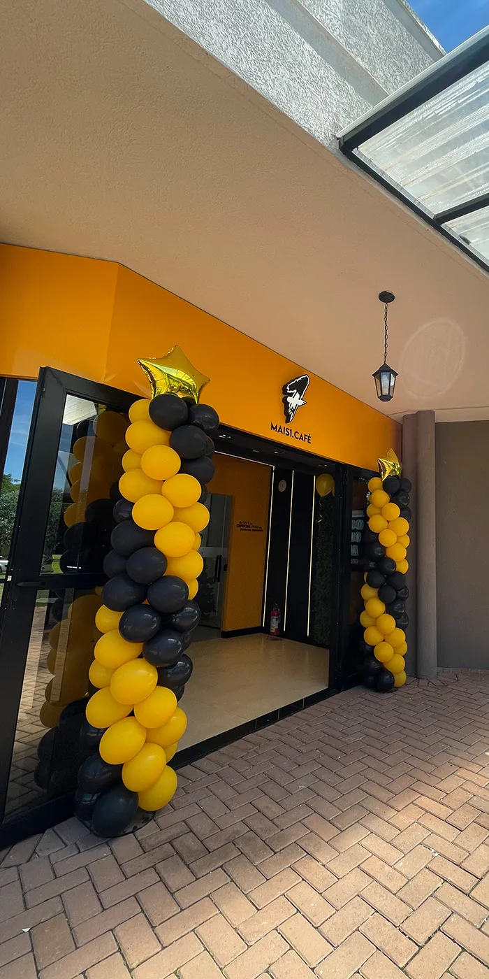 Fachada do Mais Café, com balões amarelos e pretos nas laterais da entrada e uma placa pendurada.