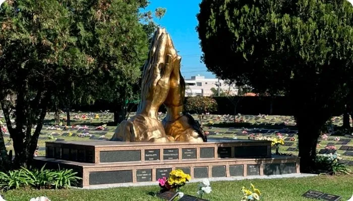 Estátua dourada de mãos em oração em um cemitério, cercada por túmulos e árvores verdes.