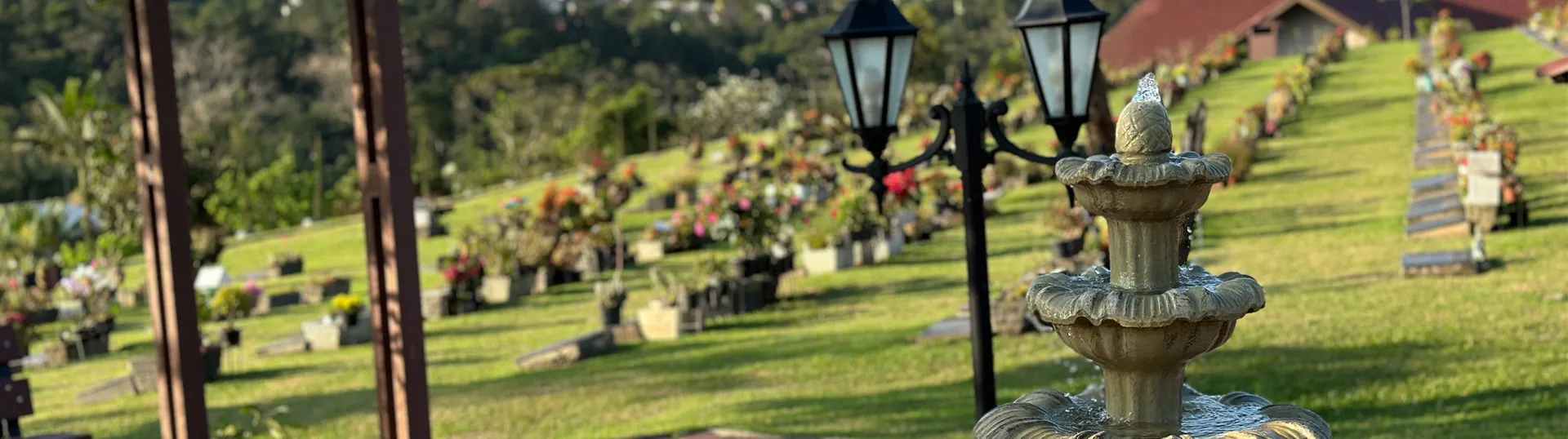 Fonte de água em um cemitério, com túmulos e flores ao fundo, cercados por uma paisagem verde.